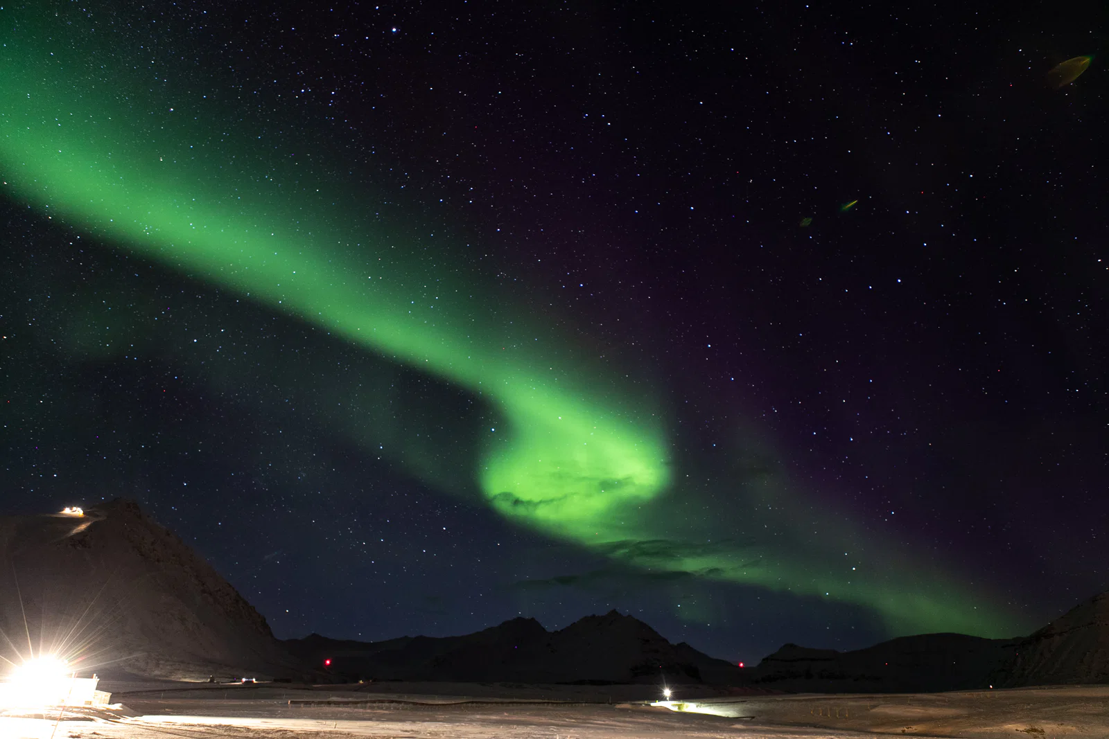 A green auroral ribbon curls across a star-filled sky above snowy mountains and buildings.