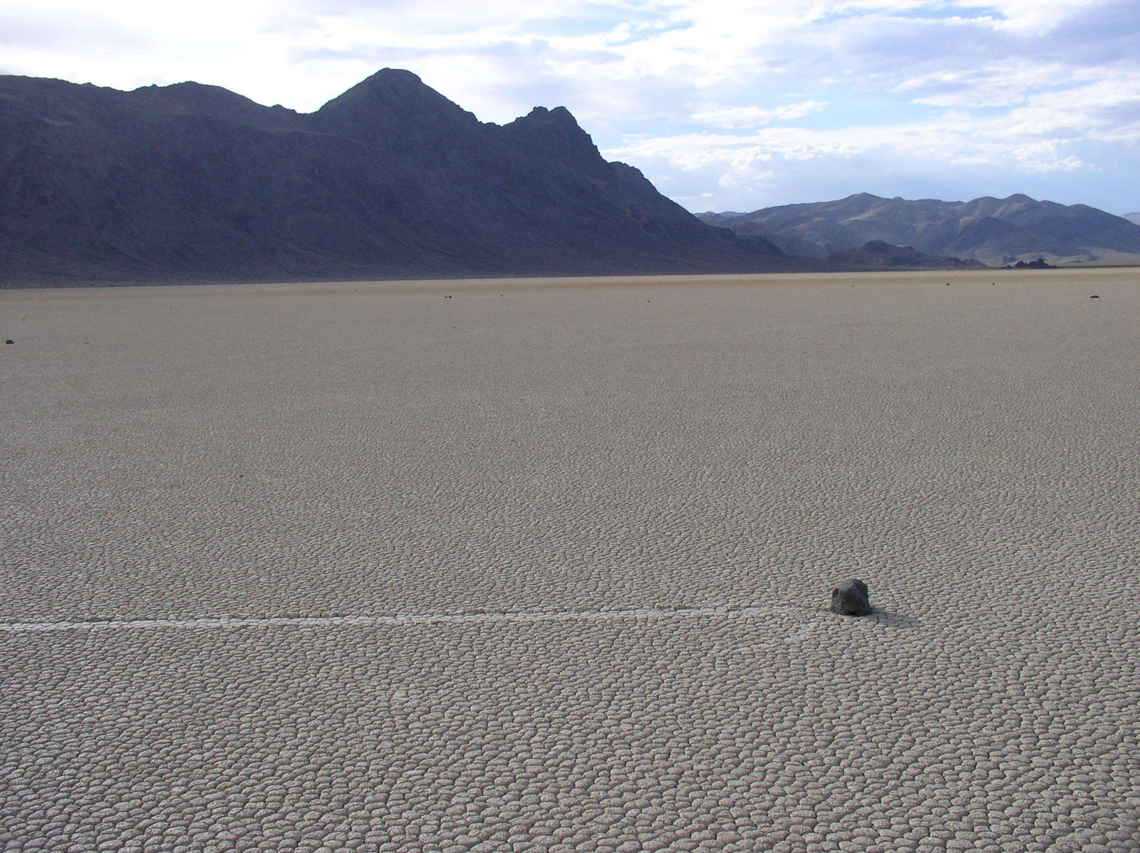 A small rock at the end of a long track across a dry lakebed, with mountains beyond.