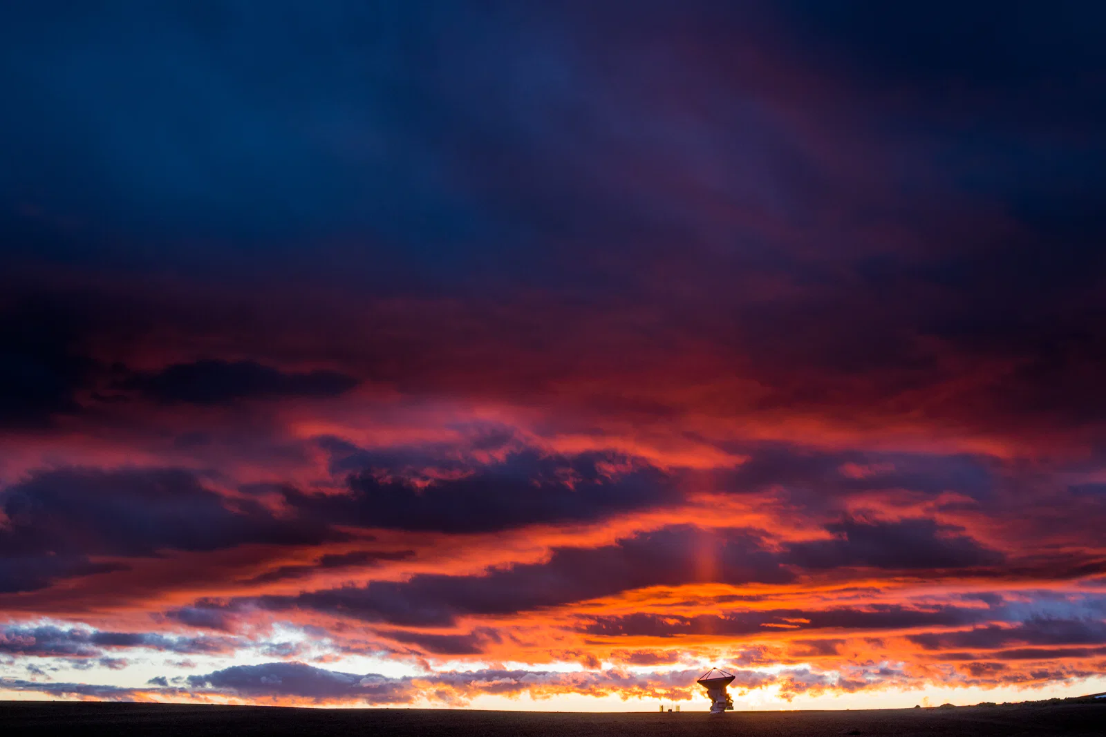 A narrow orange shaft of light rises above the sunset behind a radio-telescope dish beneath dark red clouds.