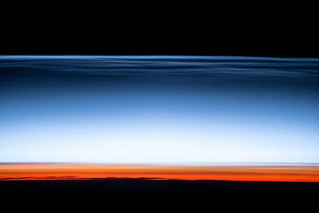 Thin blue noctilucent clouds stretch above Earth's orange-lit horizon, photographed from the International Space Station.